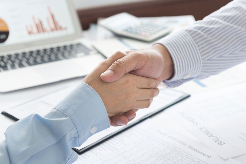 Zwei Hände schütteln sich fest über einem Schreibtisch, der mit Finanzdokumenten und einem Laptop mit Diagrammen vollgestopft ist, die eine Geschäftsvereinbarung symbolisieren.